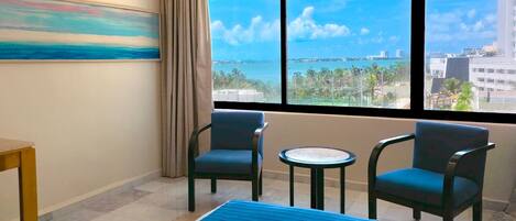 A hotel room with a bed, two chairs, a small table, a painting, and a large window with a city view.