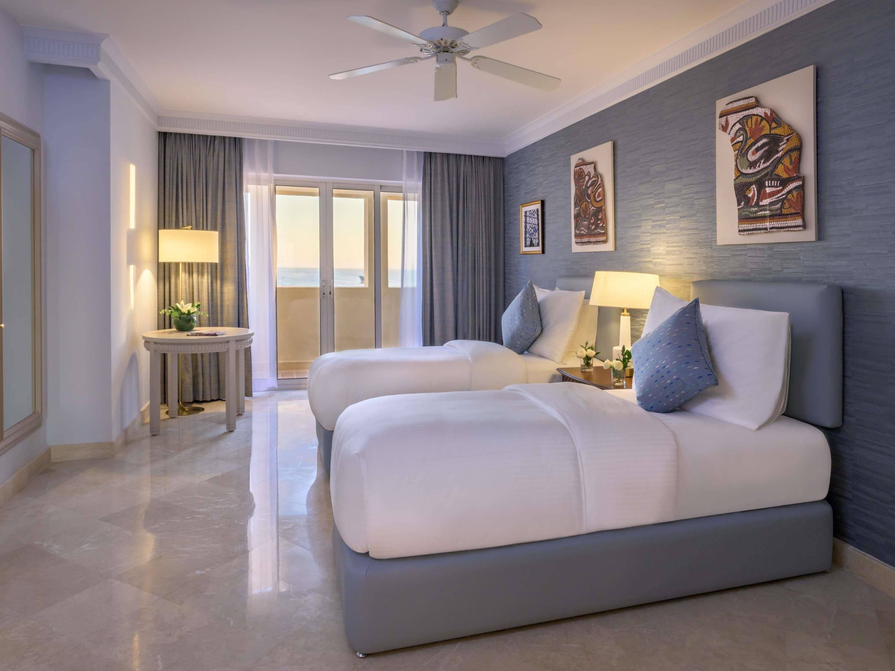 Une chambre d’hôtel avec deux lits, un ventilateur de plafond, une vue sur la mer et des œuvres d’art accrochées aux murs.