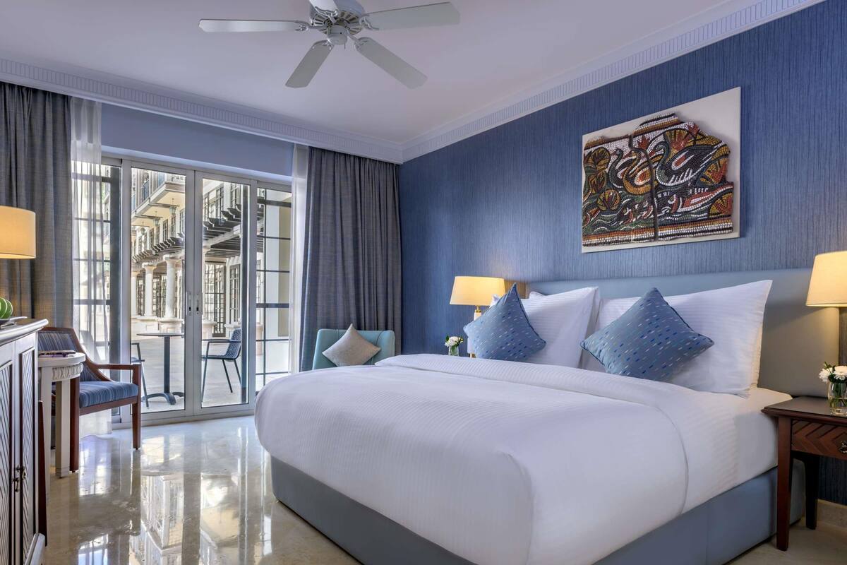 A hotel room with a large bed, two bedside lamps, a ceiling fan, a large window with curtains, and a decorative wall piece.