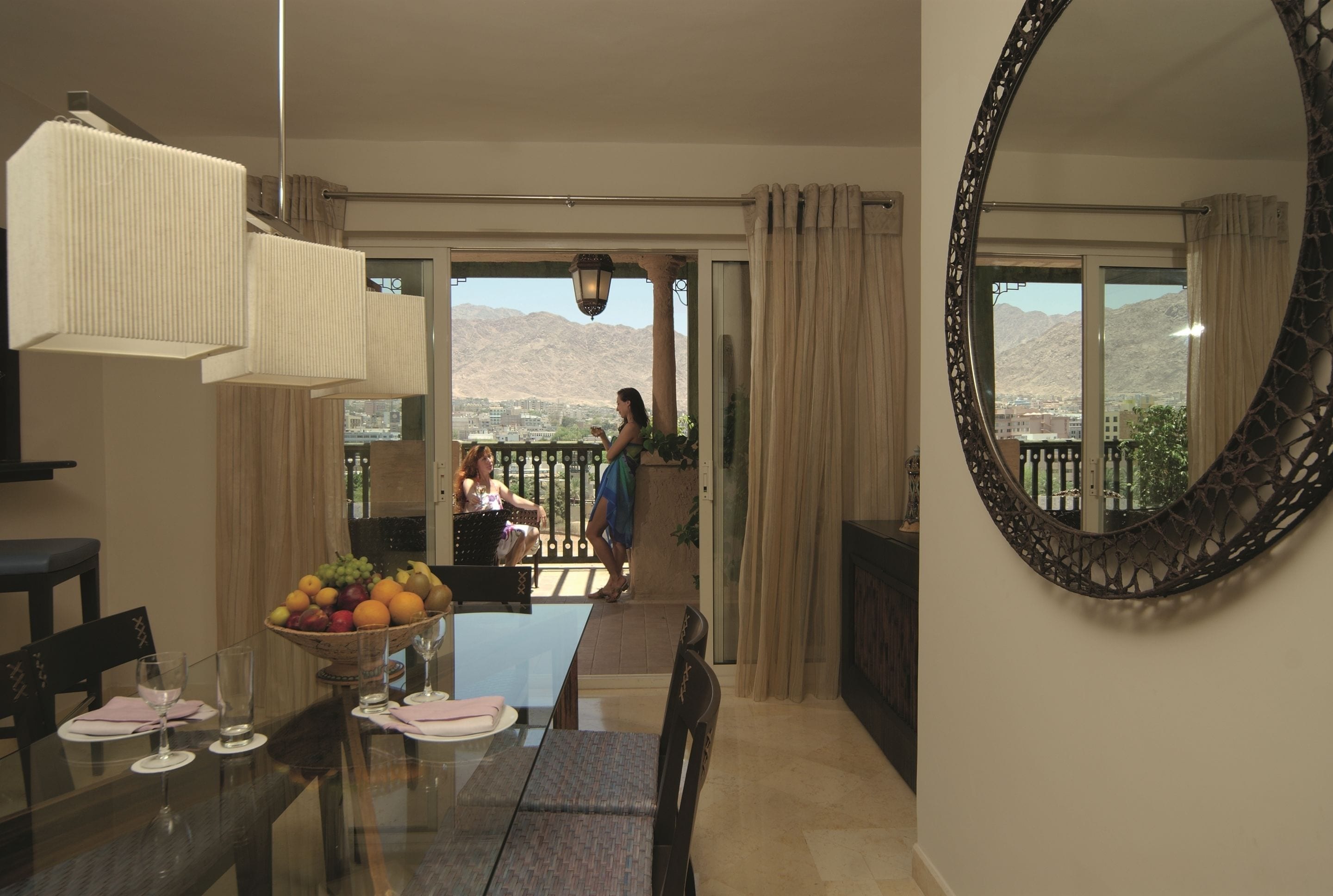 A dining area with a glass table, chairs, and a fruit bowl. There is a large mirror on the wall and a view of mountains through the windows.