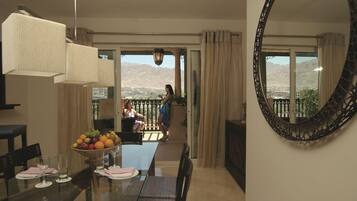 A dining area with a glass table, chairs, and a fruit bowl. There is a large mirror on the wall and a view of mountains through the windows.