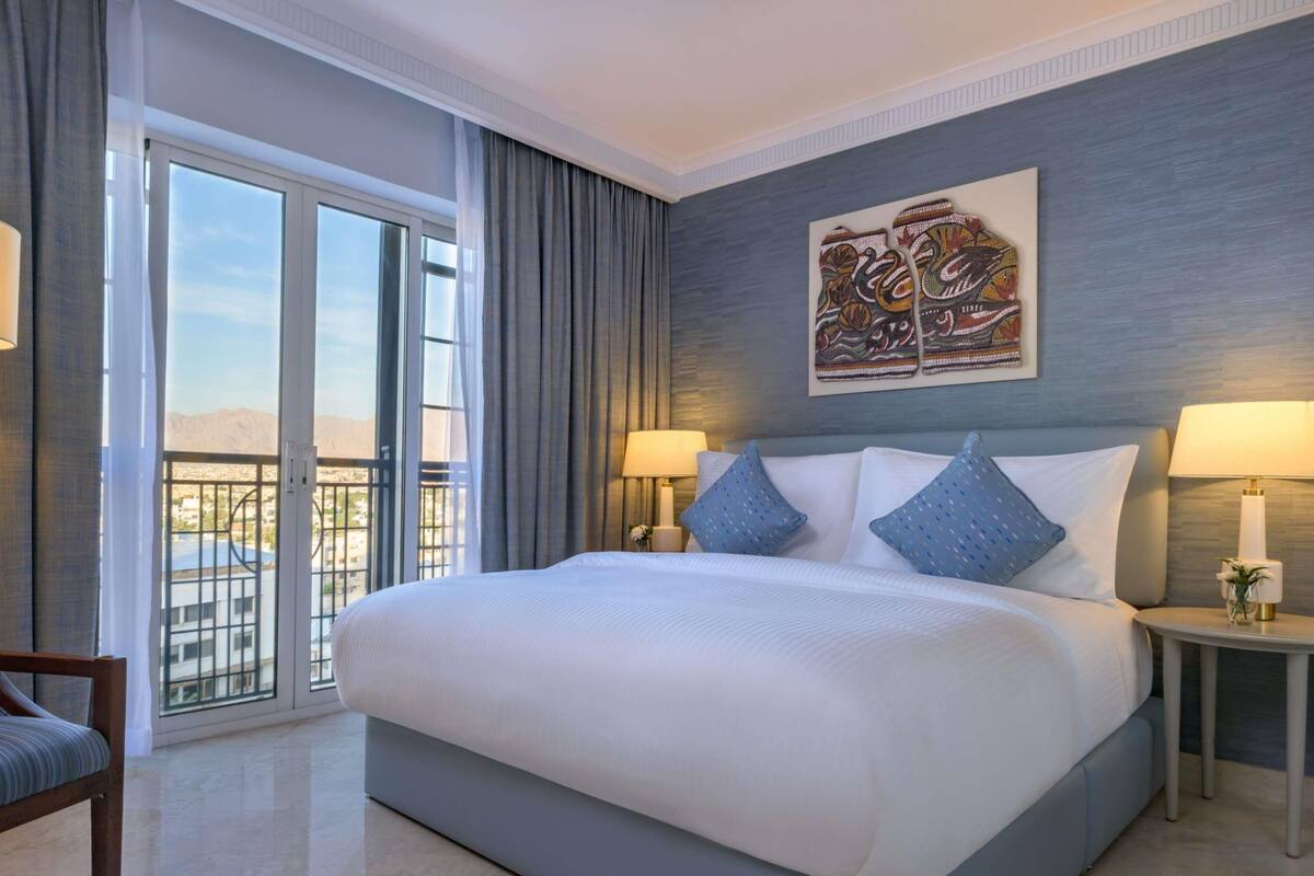 A hotel room with a large bed, two bedside lamps, a chair, a balcony with a view, and a decorative wall piece.