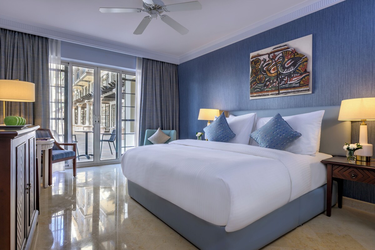 A hotel room with a large bed, two bedside tables with lamps, a ceiling fan, a large window with curtains, and a wall-mounted artwork.