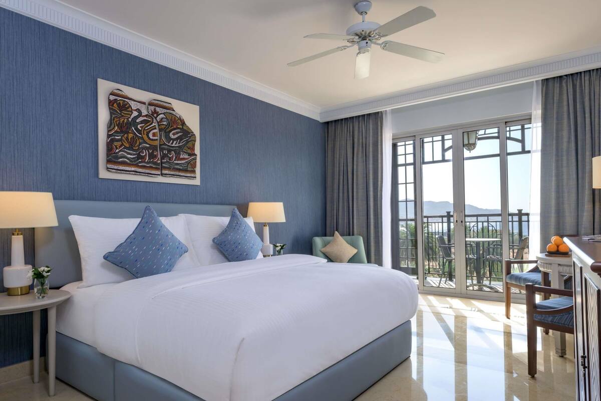 A hotel room with a bed, two bedside tables with lamps, a ceiling fan, a balcony with a table and chairs, and a large window.