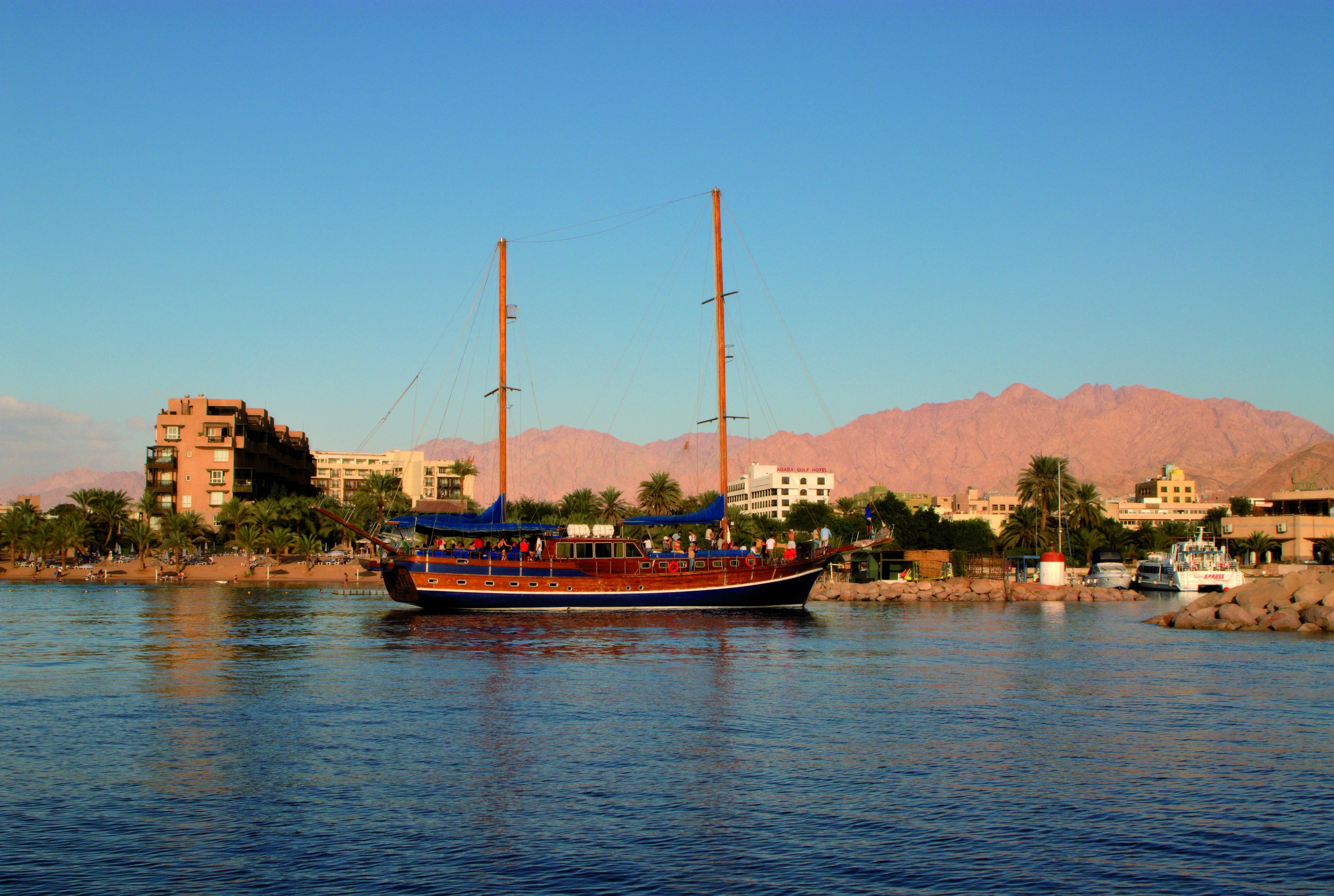 A boat with passengers on a calm river, surrounded by buildings and palm trees.