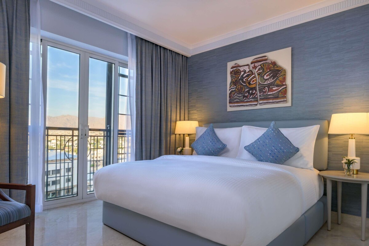 A hotel room with a large bed, two bedside lamps, a chair, a balcony with a view, and a decorative wall piece.