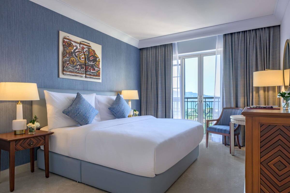 A hotel room with a large bed, two bedside tables with lamps, a chair, a desk, a large window with curtains, and a painting on the wall.