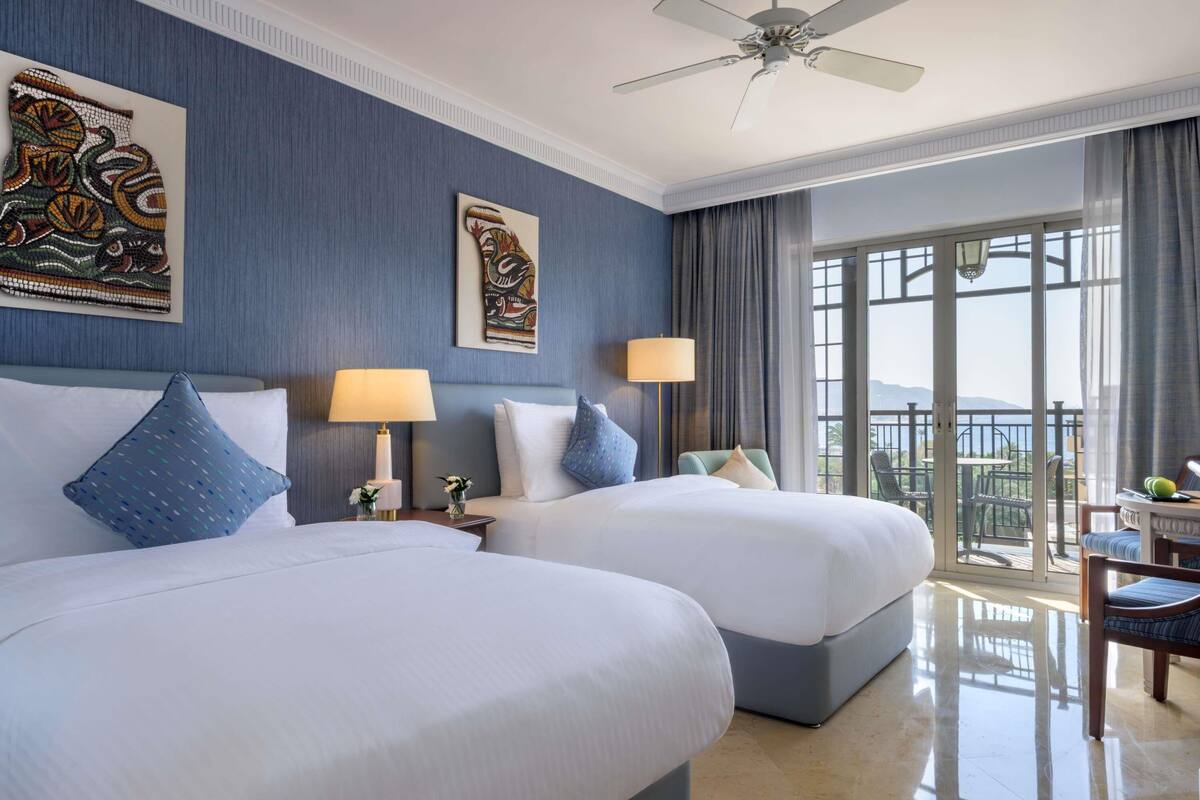 A hotel room with two beds, a ceiling fan, artwork on the walls, a balcony with a view, and a dining area.