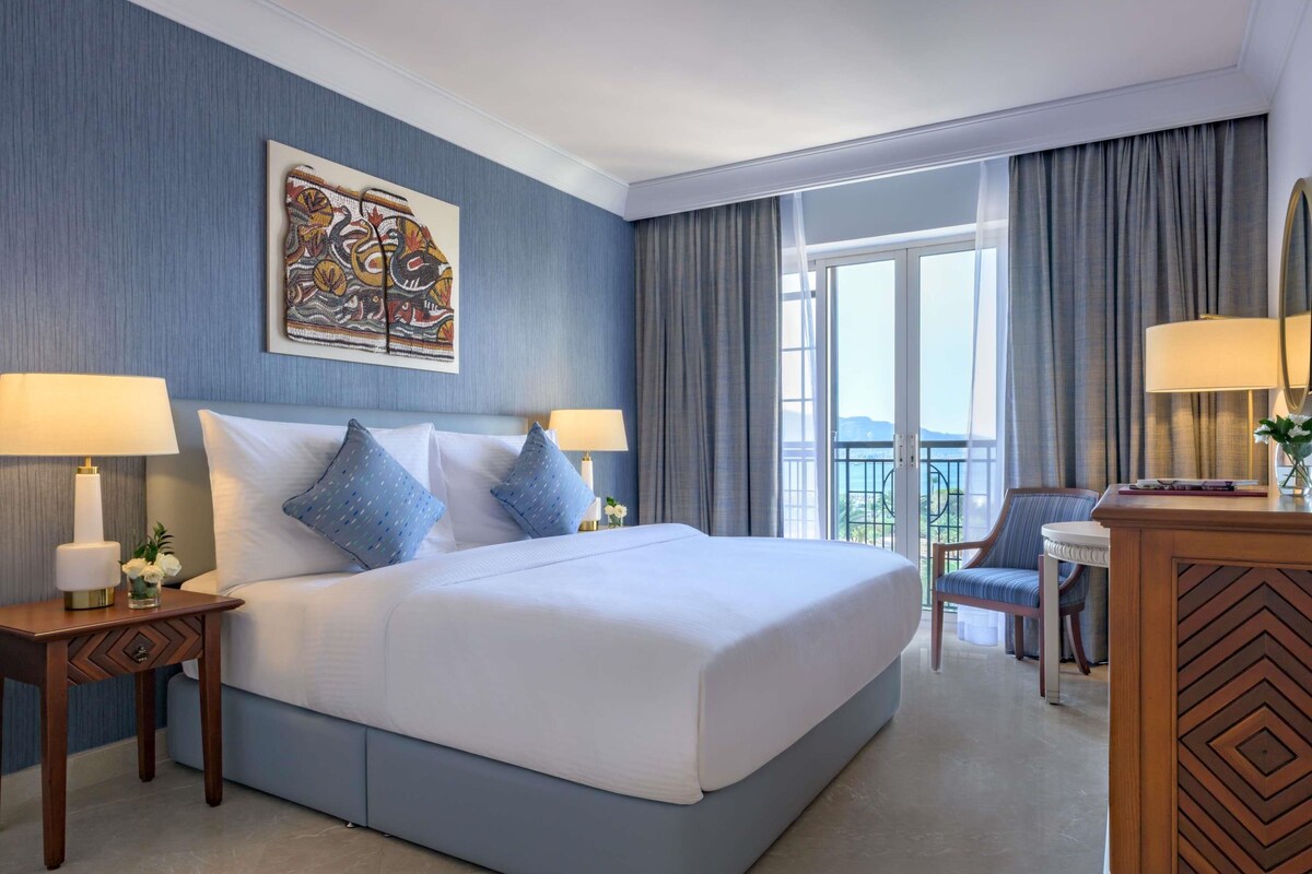 A hotel room with a large bed, two bedside tables with lamps, a chair, a desk, a large window with curtains, and a painting on the wall.