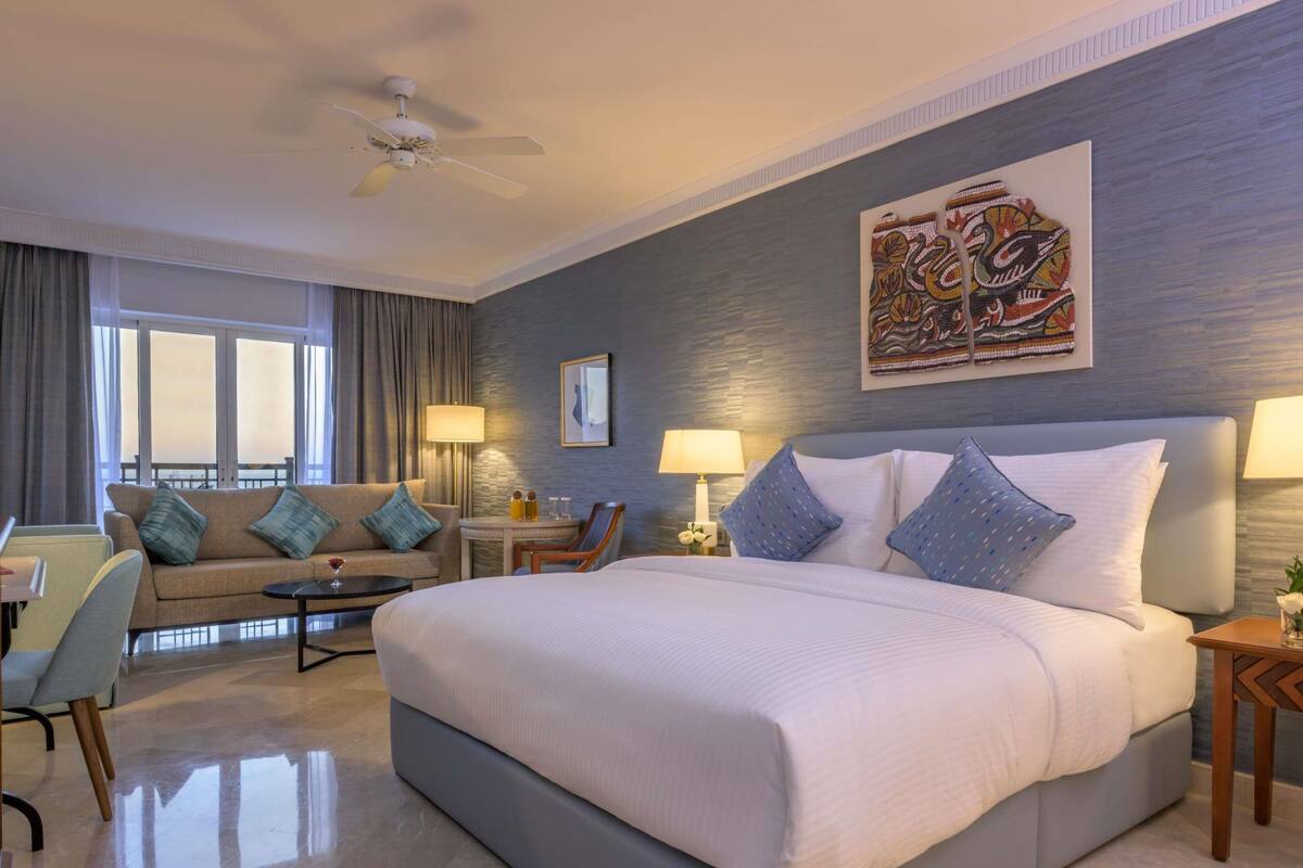 A hotel room with a large bed, a sofa, a desk, and a chair. There is a ceiling fan, a painting on the wall, and a window with curtains.