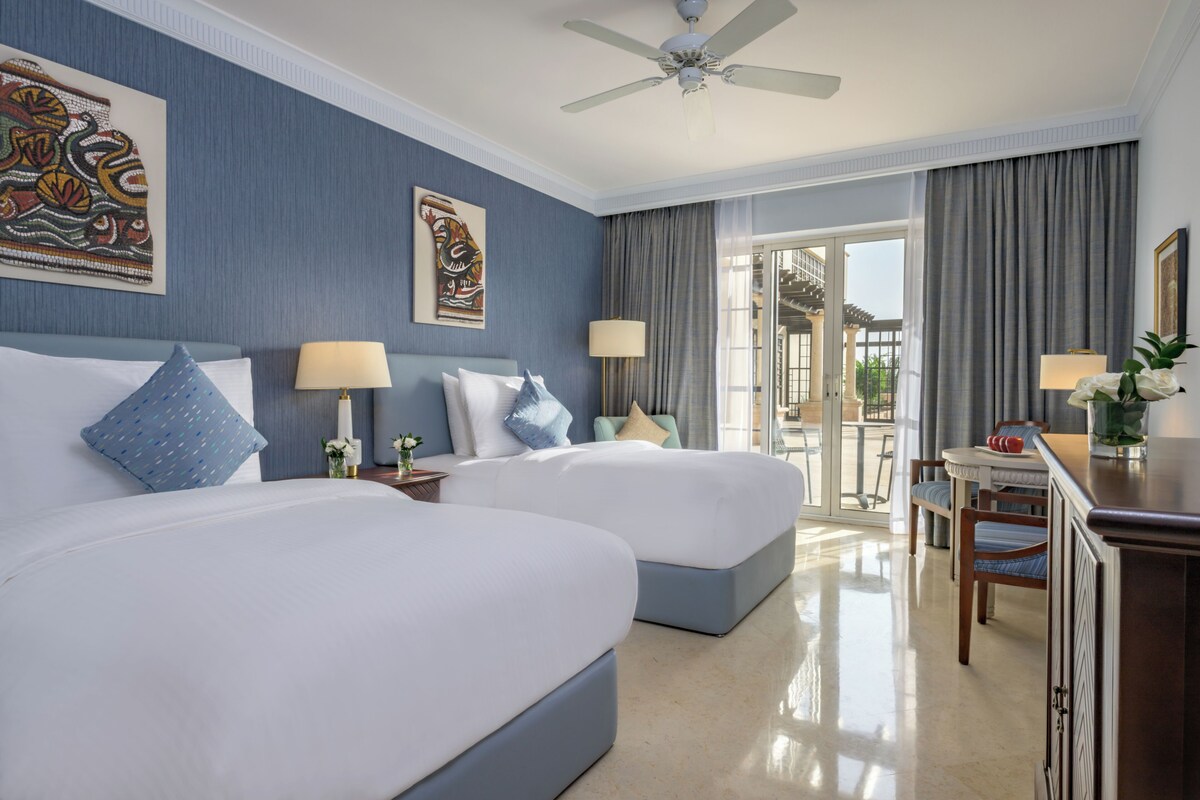 A hotel room with two beds, a desk, a chair, and a balcony with a view.