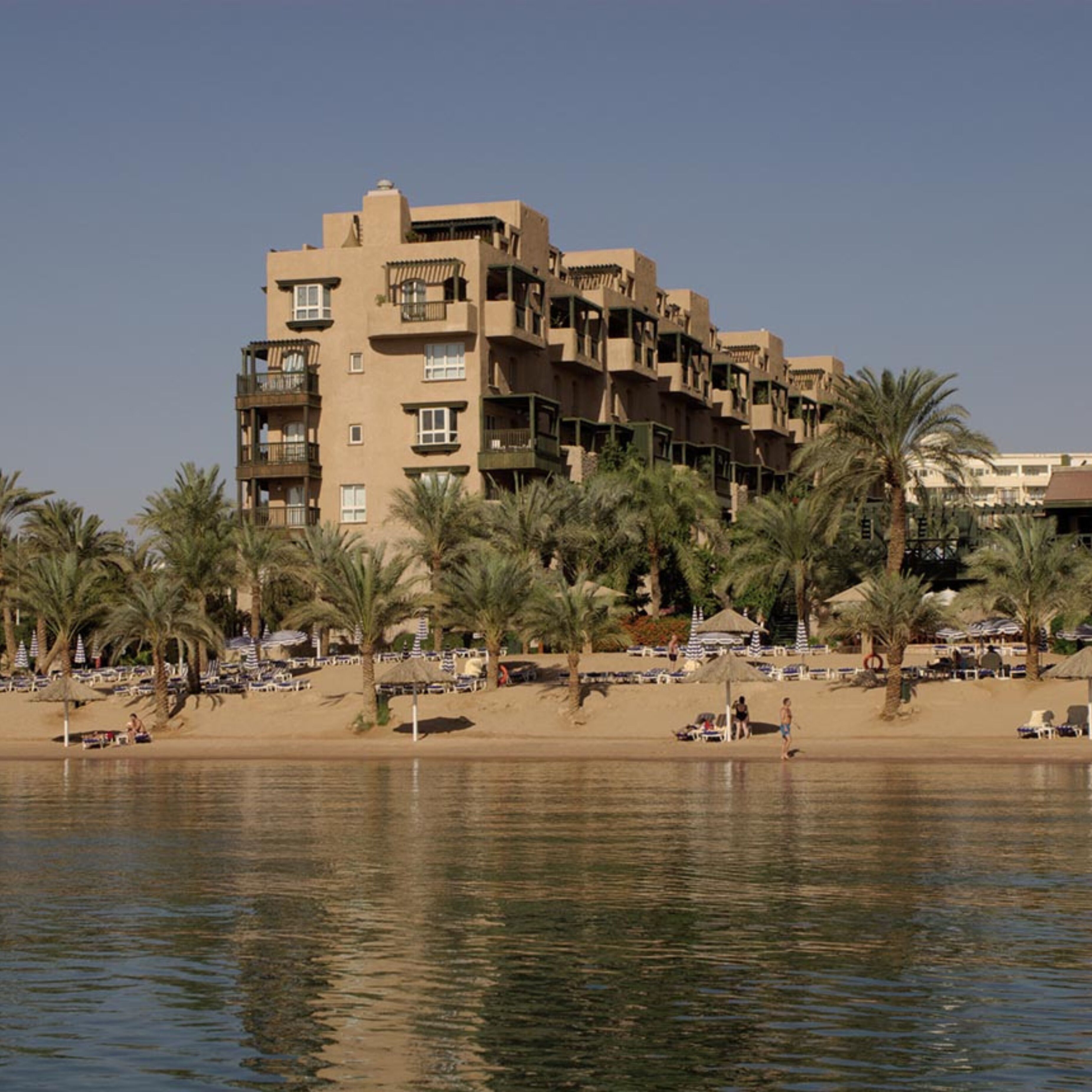 Un hotel de varios pisos con balcones, una playa arenosa al frente y palmeras.