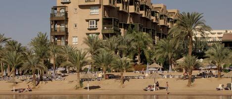 Un hotel de varios pisos con balcones, una playa arenosa al frente y palmeras.