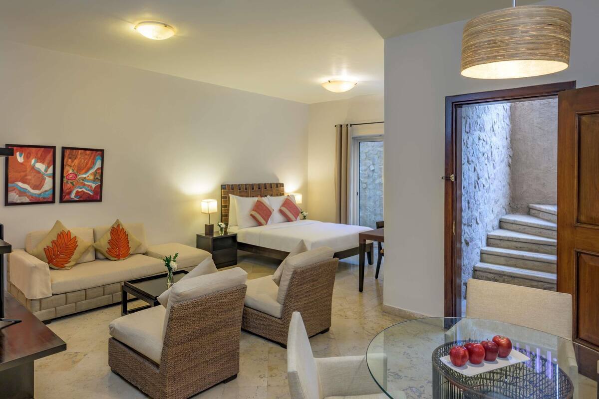 A hotel room with a bed, a sofa, two armchairs, a glass table with apples, and a staircase leading to an outdoor area.
