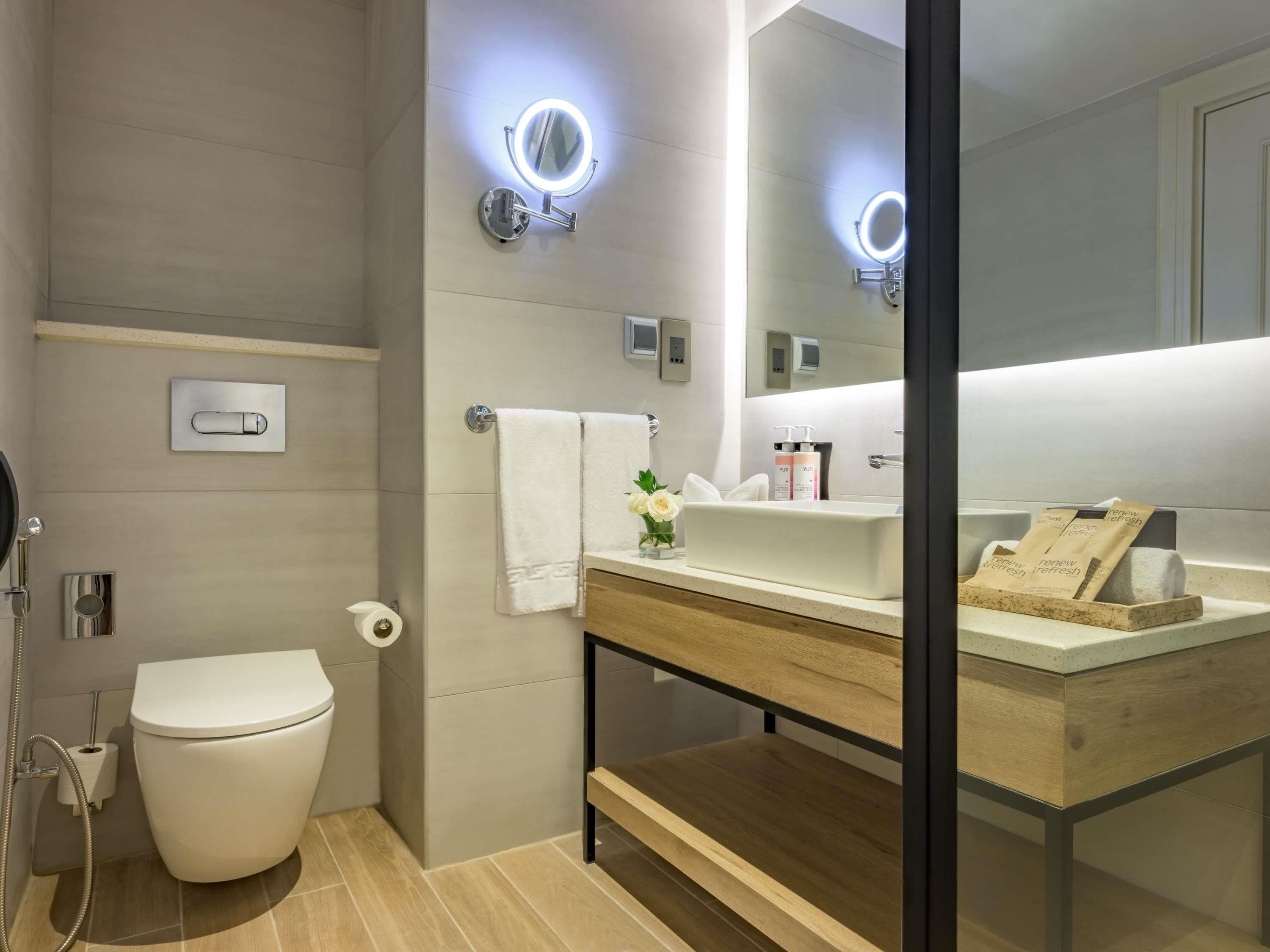 A modern bathroom with a toilet, sink, and mirror with lights, a towel rack, and a small plant.