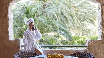 A person in a white bathrobe standing by a glass table with a bowl of lemons, a wicker chair, and a view of palm trees through a stone archway.