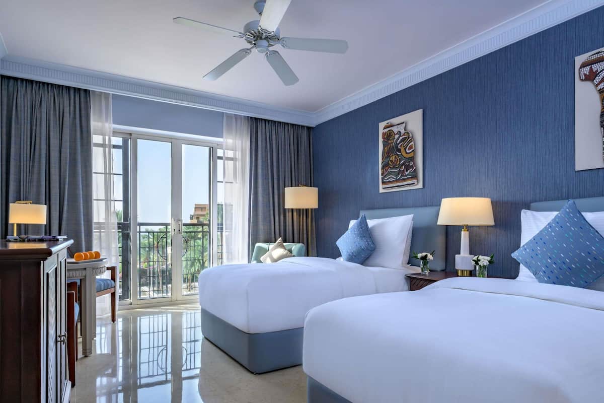 A hotel room with two beds, a ceiling fan, a balcony with a view, and a desk with a lamp.