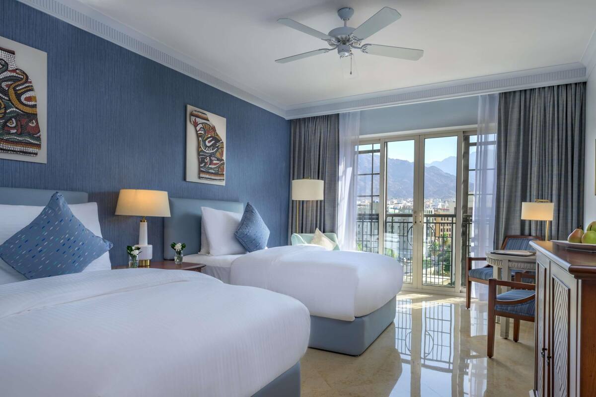 A hotel room with two beds, a ceiling fan, a balcony with a view, and a desk with a lamp.