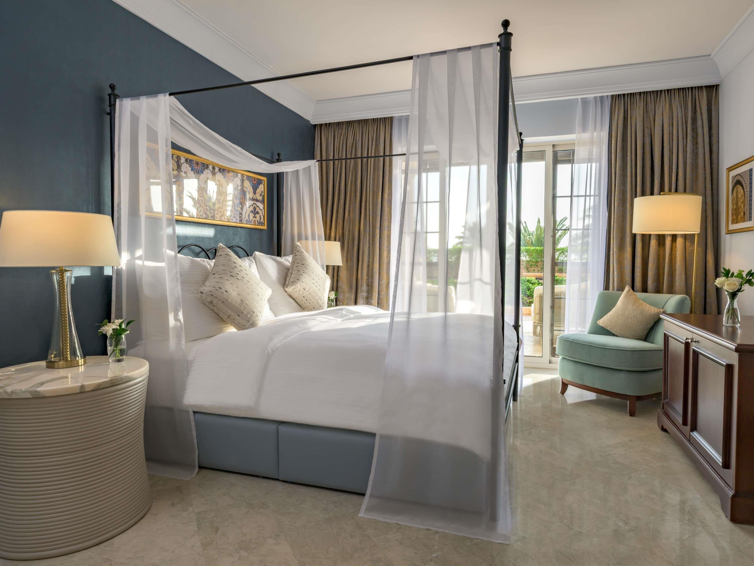 A hotel room with a four-poster bed, a round side table, a chair, and a desk with a lamp.