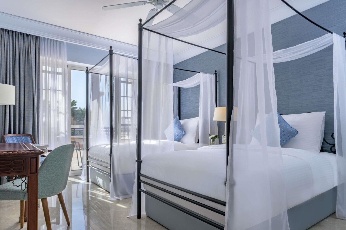 A four-poster bed with sheer curtains, a desk with a chair, and a view of the outdoors.