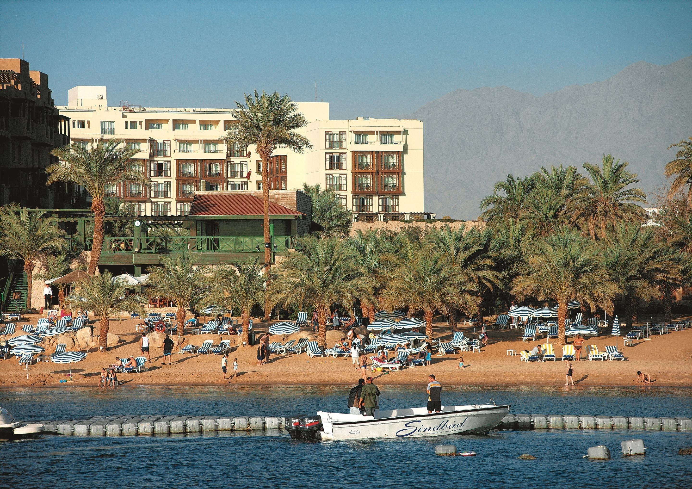 A beachfront hotel with a pool area, palm trees, and a boat docked by the water.
