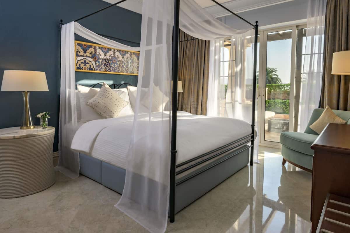A four-poster bed with sheer curtains, a nightstand with a lamp, a vase with flowers, and a view of the outdoors through a sliding glass door.