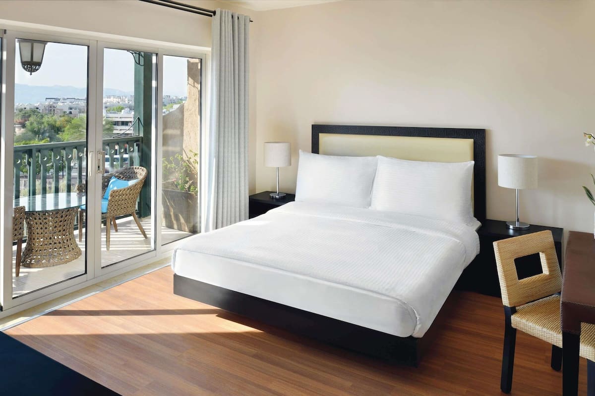 A hotel room with a large bed, two bedside tables with lamps, a wicker chair, and a balcony with a view of the city.