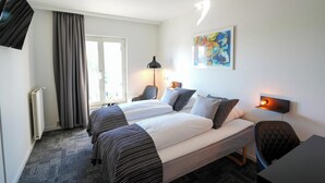 A hotel room with two beds, a desk, a chair, a radiator, a window with curtains, a painting on the wall, and a wall-mounted lamp. - Østergaards Hotel (Herning)
