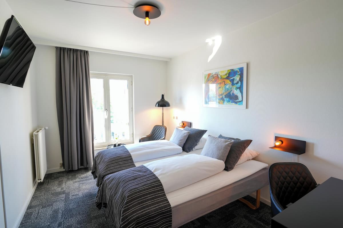 Une chambre d’hôtel avec deux lits, un bureau, une chaise, un radiateur, une fenêtre avec des rideaux, un tableau au mur et une lampe fixée au mur.