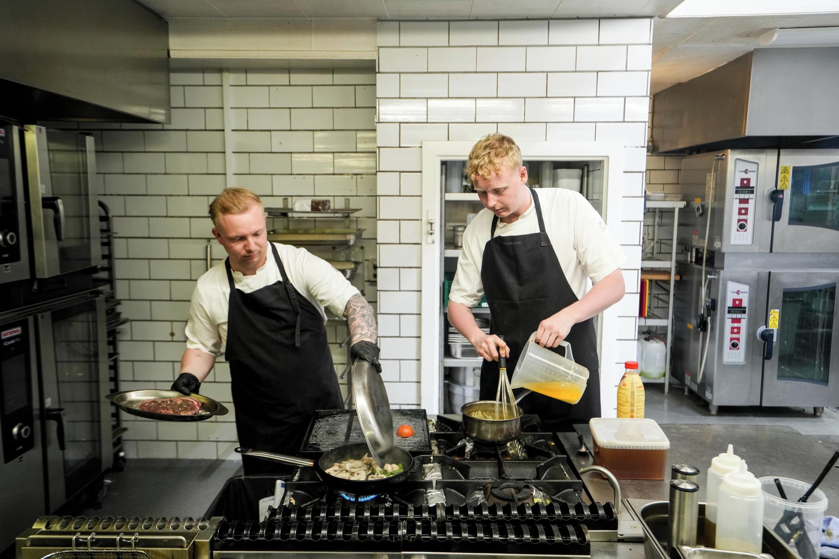 A kitchen with two chefs preparing food, stainless steel appliances, and a tiled backsplash.