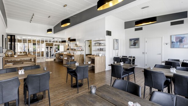 A spacious dining area with multiple tables and chairs, a kitchen area with shelves and appliances, and a large window providing natural light. - Østergaards Hotel (Herning)