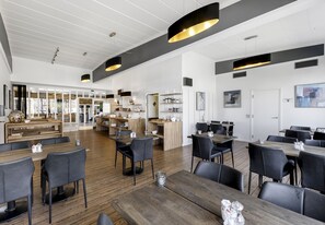 A spacious dining area with multiple tables and chairs, a kitchen area with shelves and appliances, and a large window providing natural light. - Østergaards Hotel (Herning)
