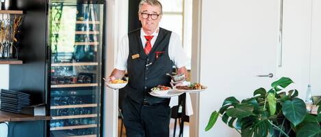 A person in a black vest and white shirt carrying trays with food and a wine cooler.