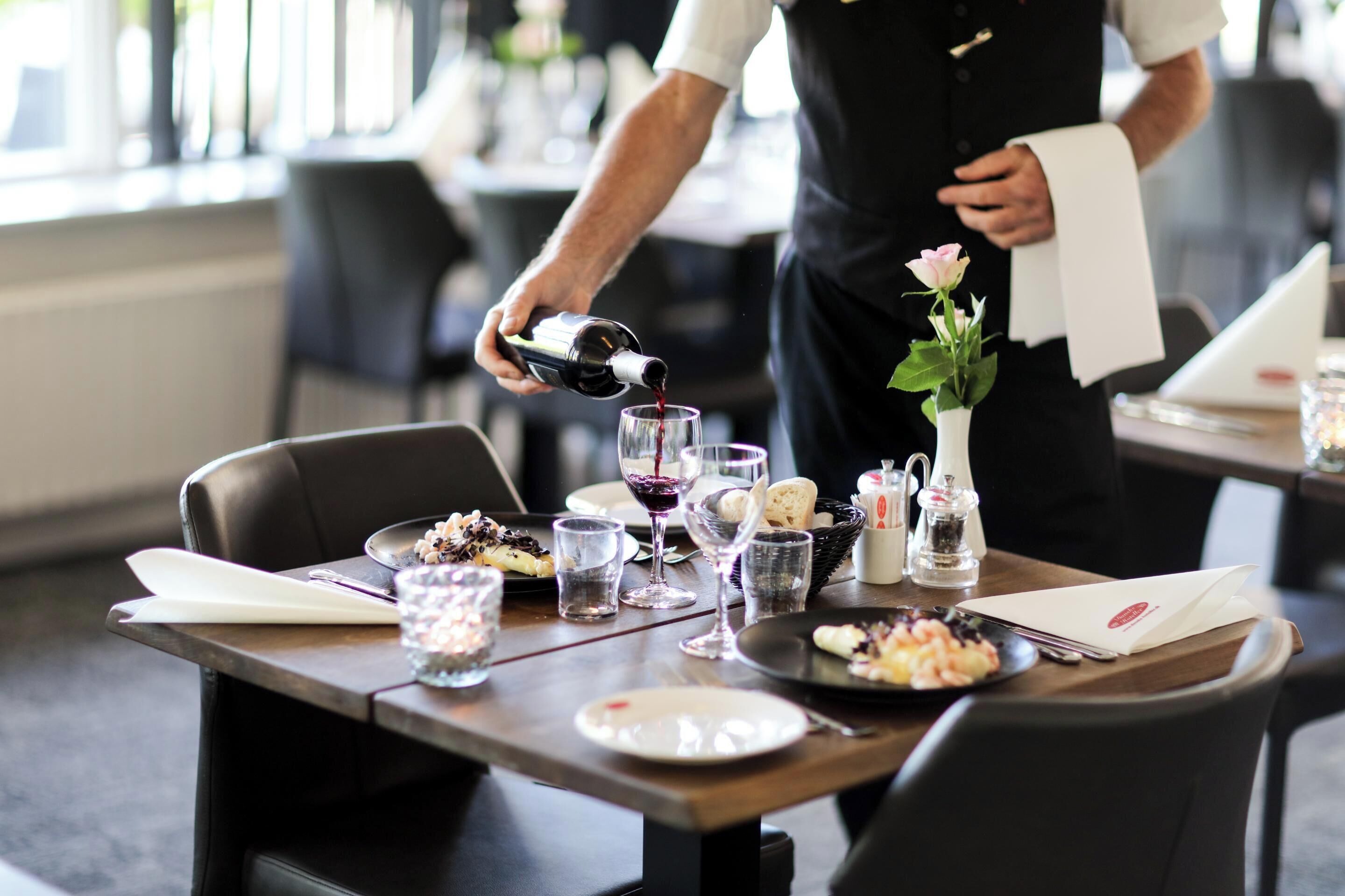 Una mesa de comedor dispuesta con platos, vasos y una botella de vino, mientras un camarero sirve vino.
