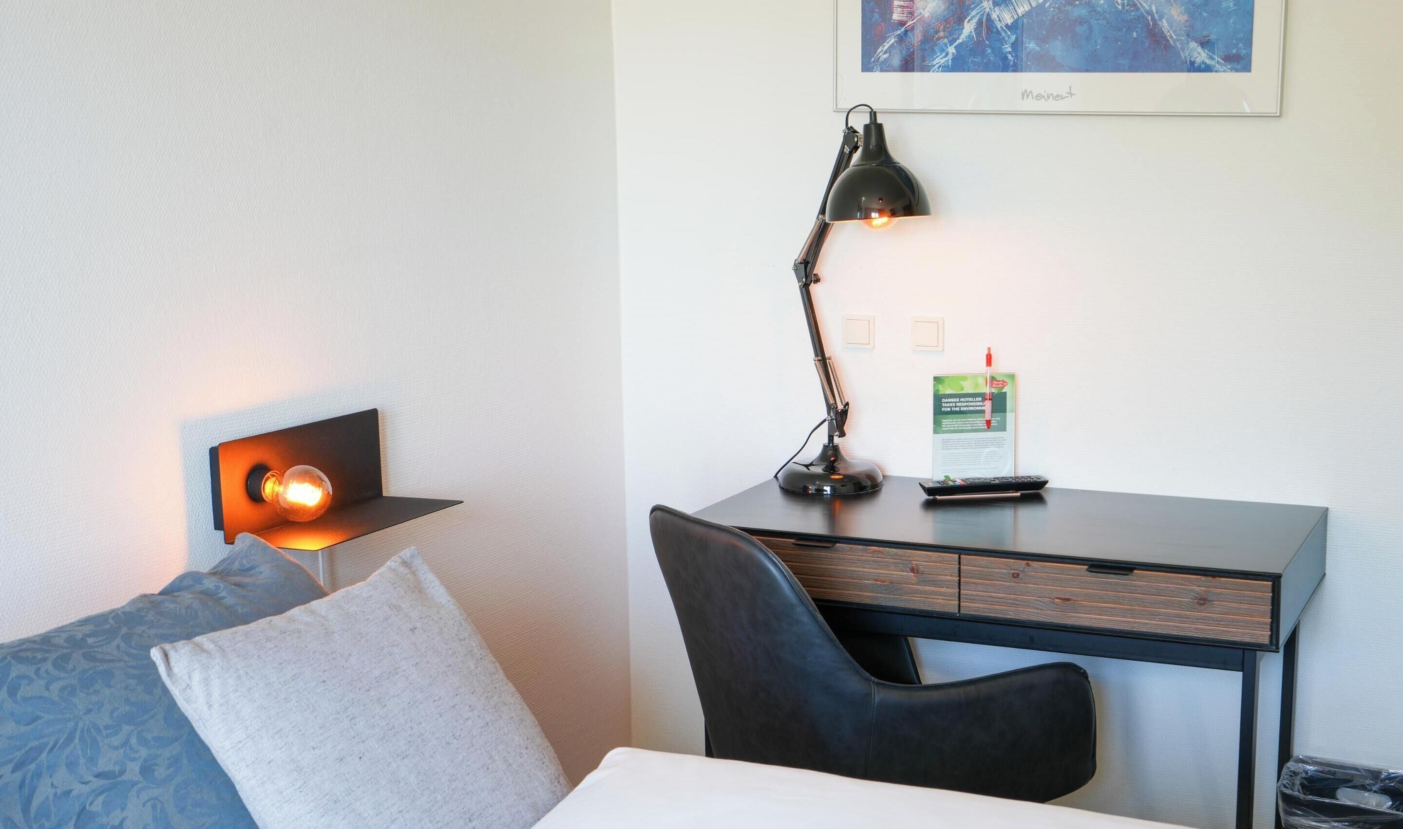A hotel room with a bed, a desk with a lamp, a chair, a framed artwork on the wall, and a brochure on the desk.