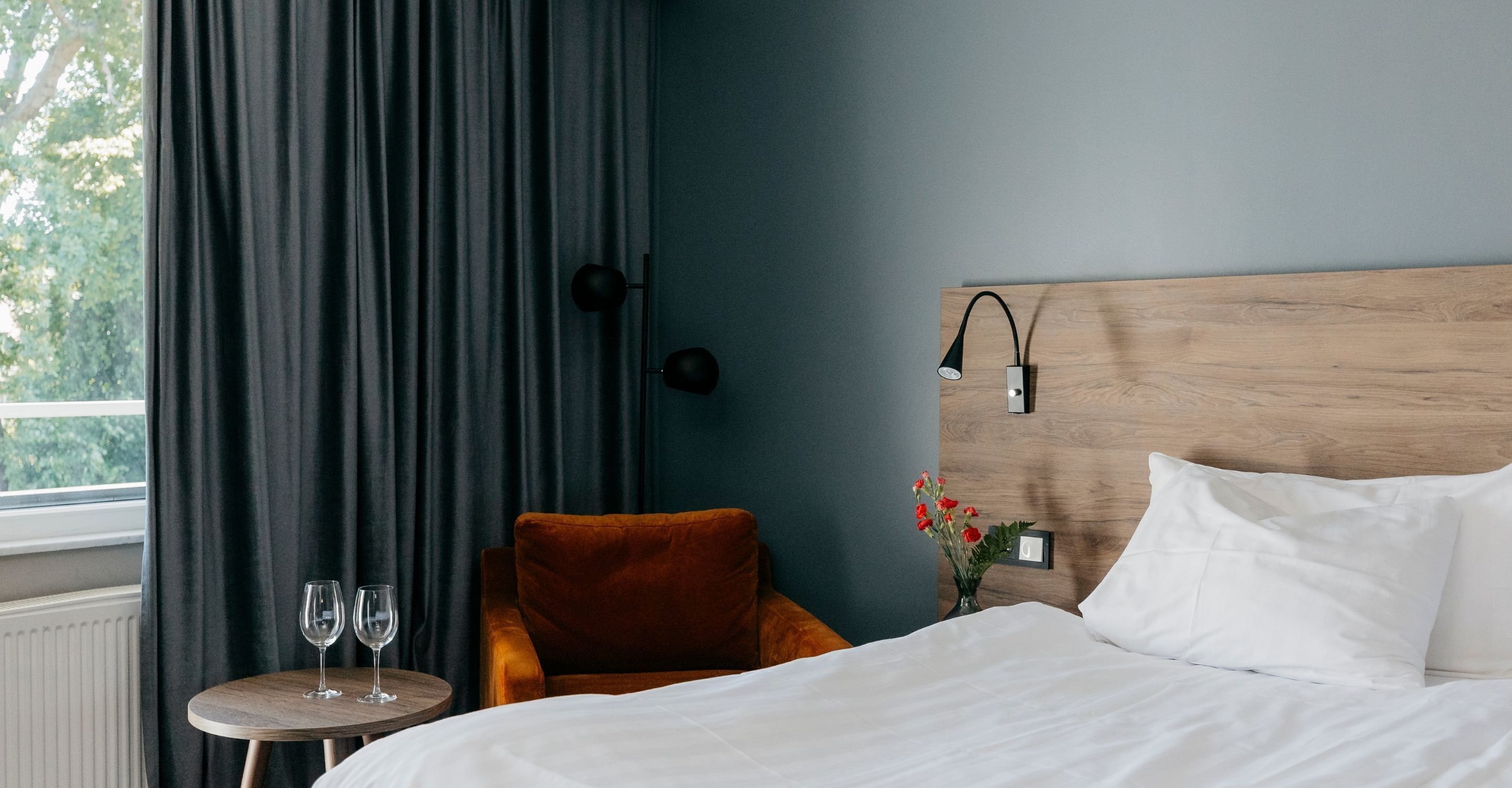 Habitación de hotel con cama, cabecero de madera, mesita de noche con l ámpara, una silla, una pequeña mesa con dos copas de vino y un jarrón con flores.