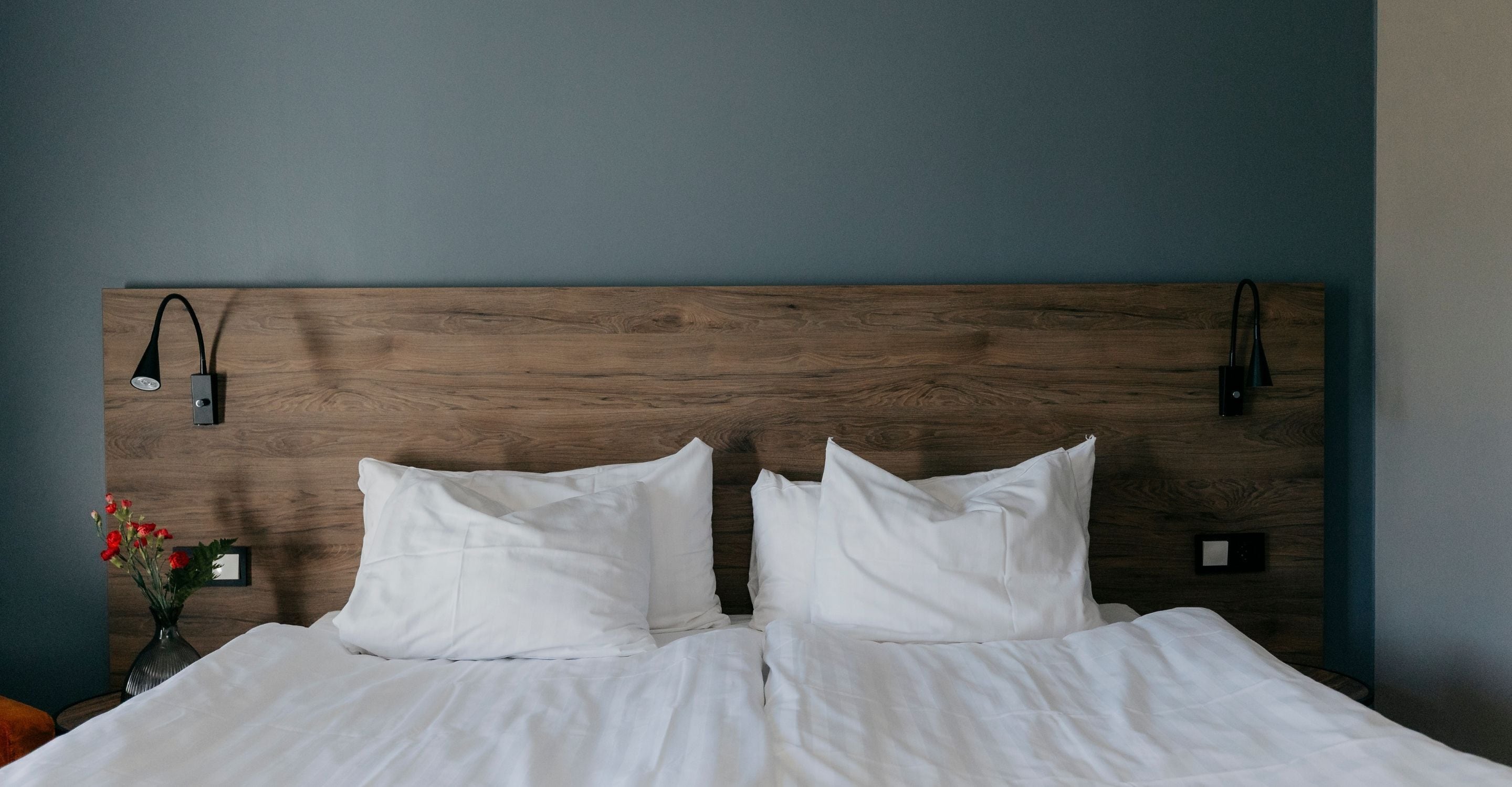 A neatly made bed with white linens, a wooden headboard, and a small bedside table with a vase of red flowers.