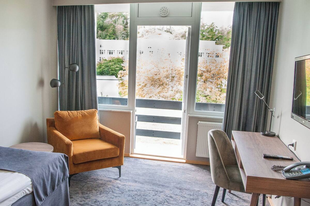 Ein Hotelzimmer mit Bett, Sofa, Schreibtisch und Blick auf ein Gebäude durch eine Glastür.
