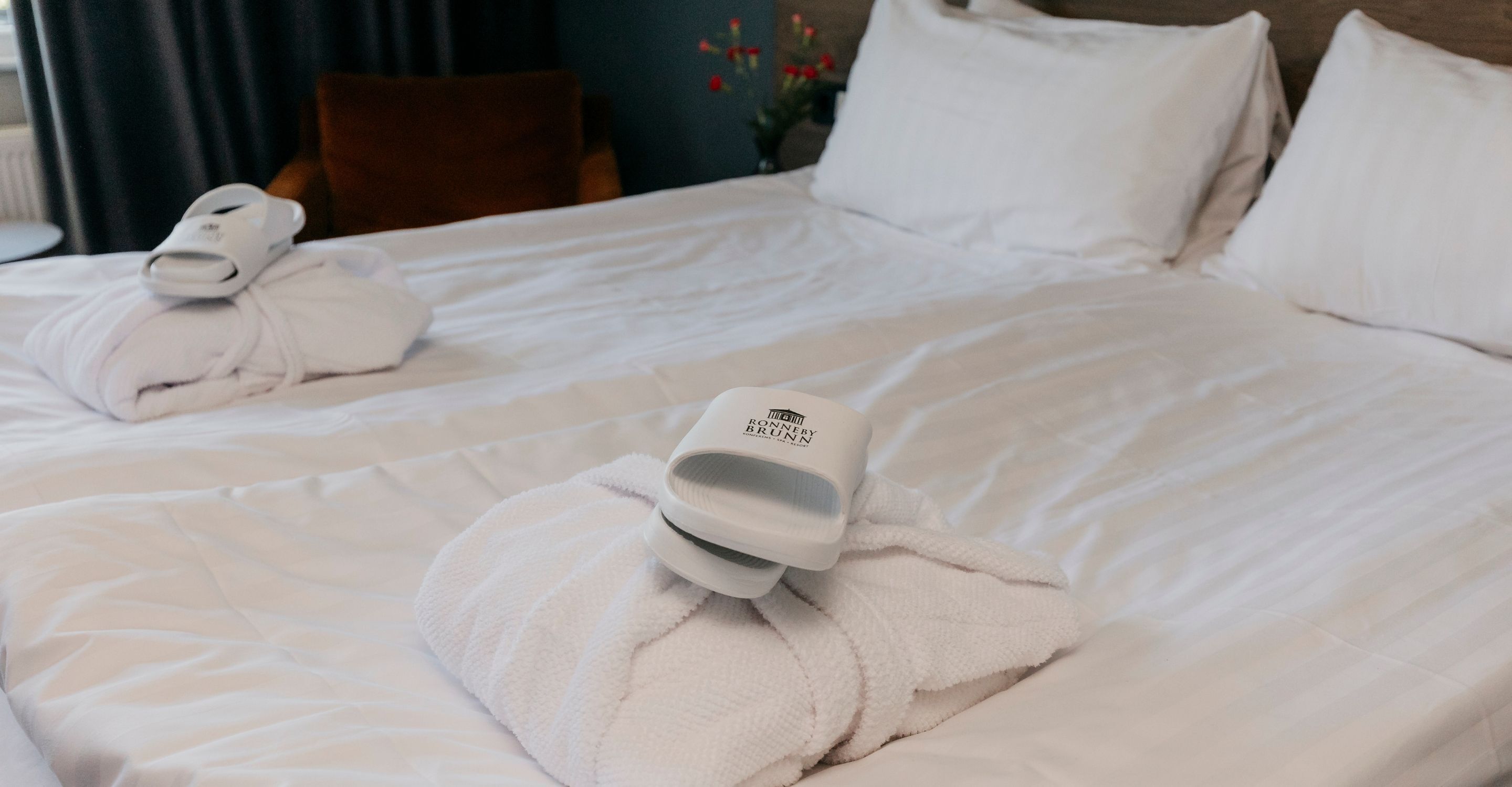 Quarto de hotel com cama arrumada, roupão pendurado ao lado e secador de cabelo sobre a cama.