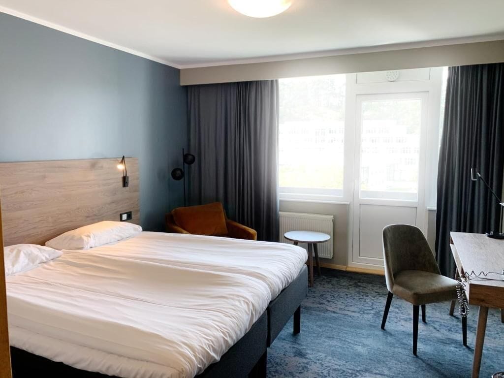 Habitación de hotel con una cama grande, una mesita redonda pequeña, una silla y un ventanal con cortinas.