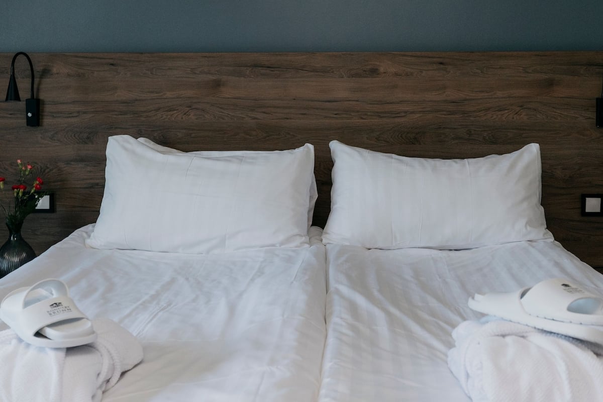 Ein Doppelbett mit weißen Leinen, zwei Kissen und je Seite zwei weiße Hausschuhe.
