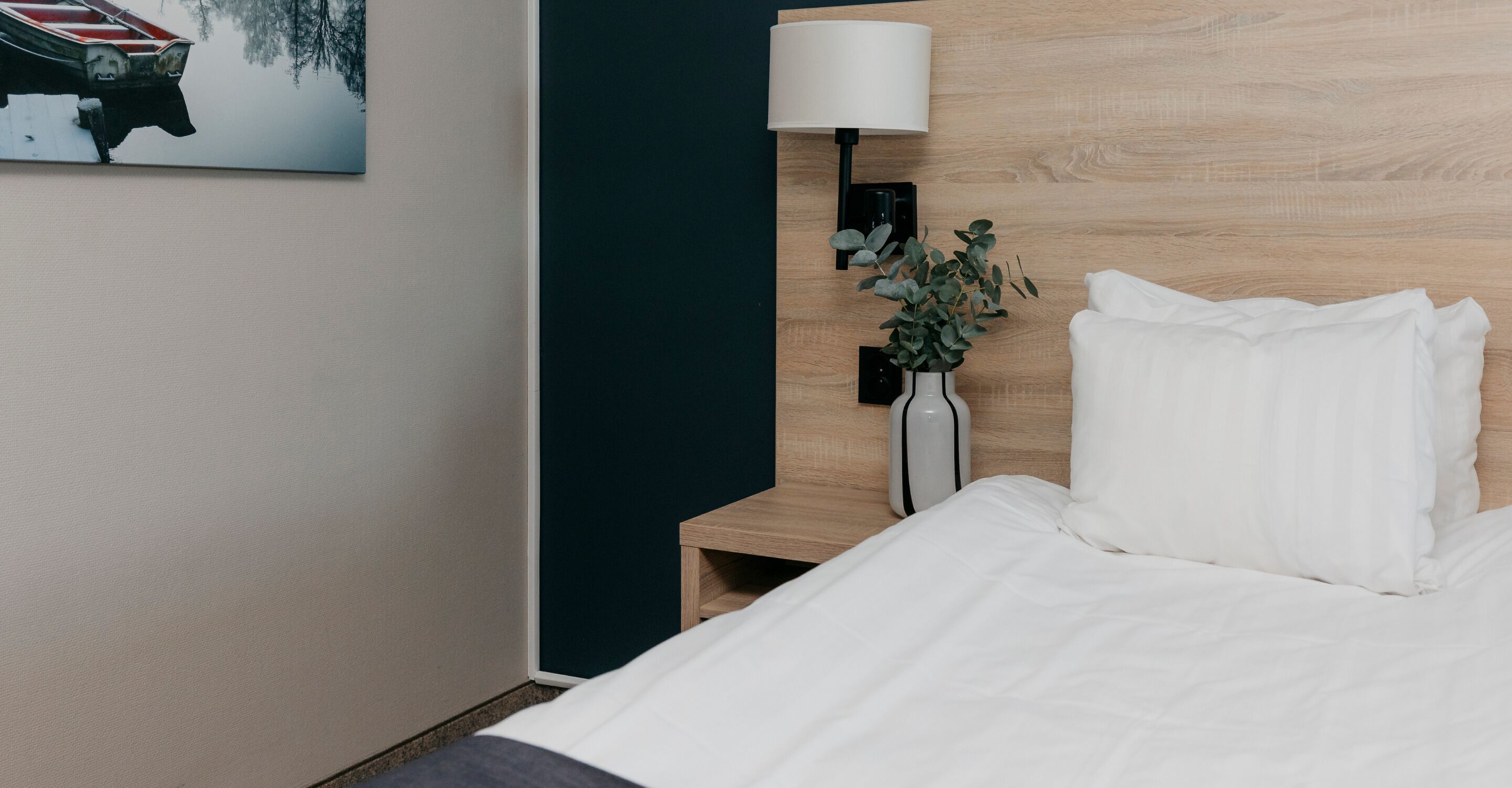 Ein ordentlich bezogenes Bett mit weißen Leinen, eine Nachttischlampe, ein kleiner Tisch mit einer Pflanzvase und ein Gemälde eines Bootes an der Wand.