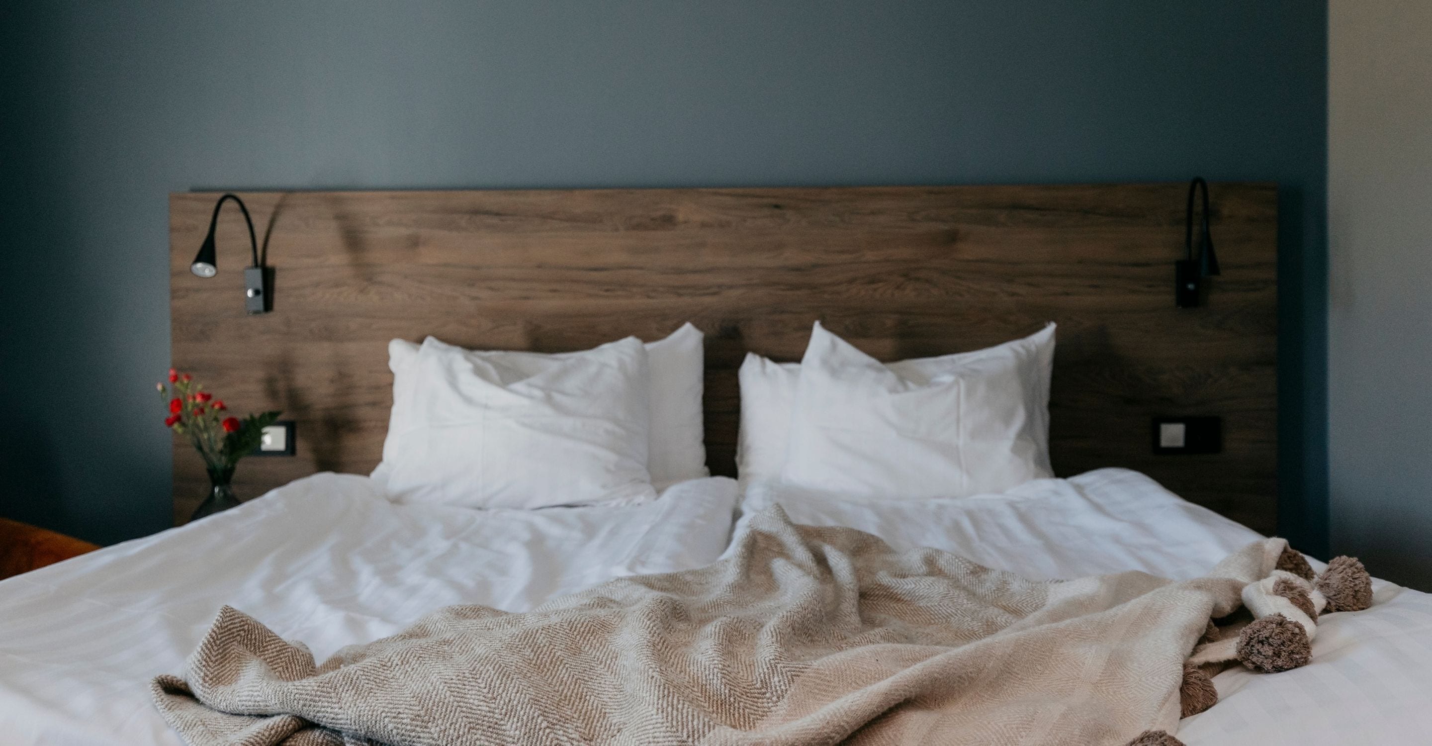 A neatly made bed with white linens and a beige blanket, a wooden headboard, and a small bedside table with a lamp.