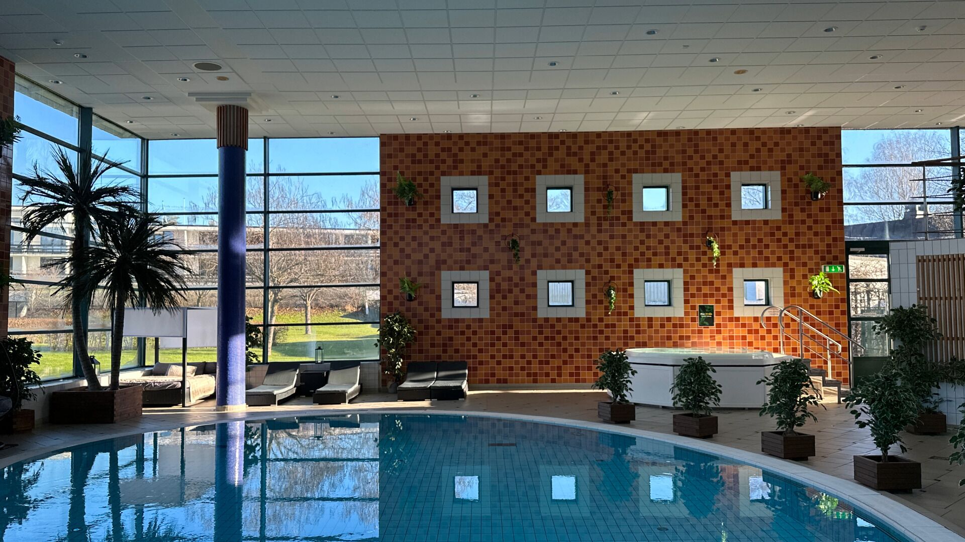 Una piscina interior con vistas a un paisaje verde a través de amplias ventanas, un muro con azulejos y pequeñas ventanas, y varias plantas en macetas.