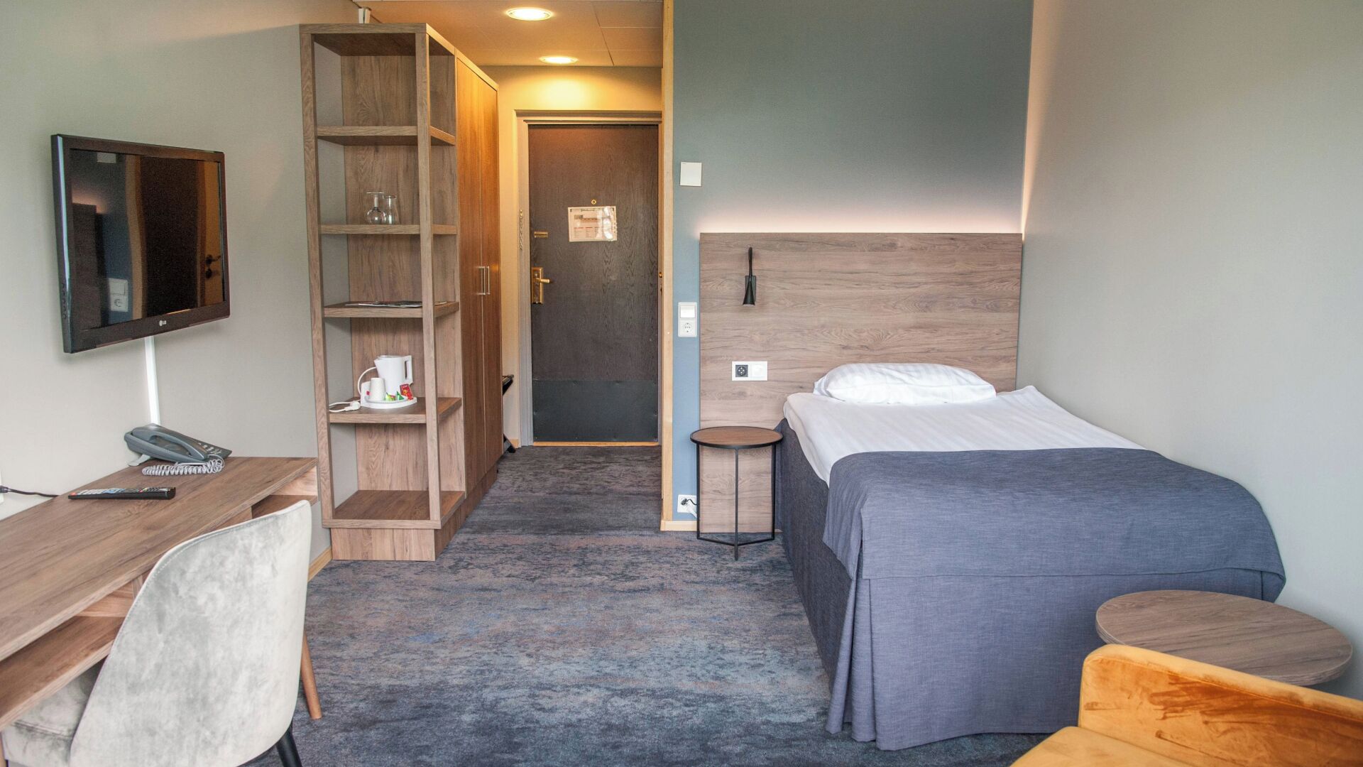 A hotel room with a bed, a desk with a chair, a TV, a wardrobe, and a small table.