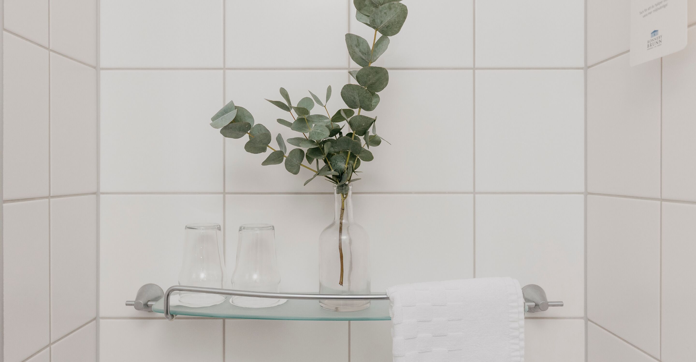 Uma casa de banho com azulejos brancos, uma prateleira de vidro com dois copos transparentes, um vaso com folhagem verde e uma toalha branca num porta-toalhas.