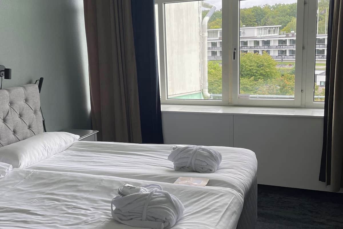 Ein Hotelzimmer mit einer geblümten Kopfteil, zwei Betten mit weißen Bettwäsche, einem Fenster mit Blick auf Gebäude und Bäume sowie einer Wandleuchte.