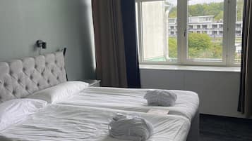 Una habitación de hotel con cabecera capitonada, dos camas con ropa blanca, un ventanal con vistas a edificios y árboles, y una lámpara fijada a la pared.