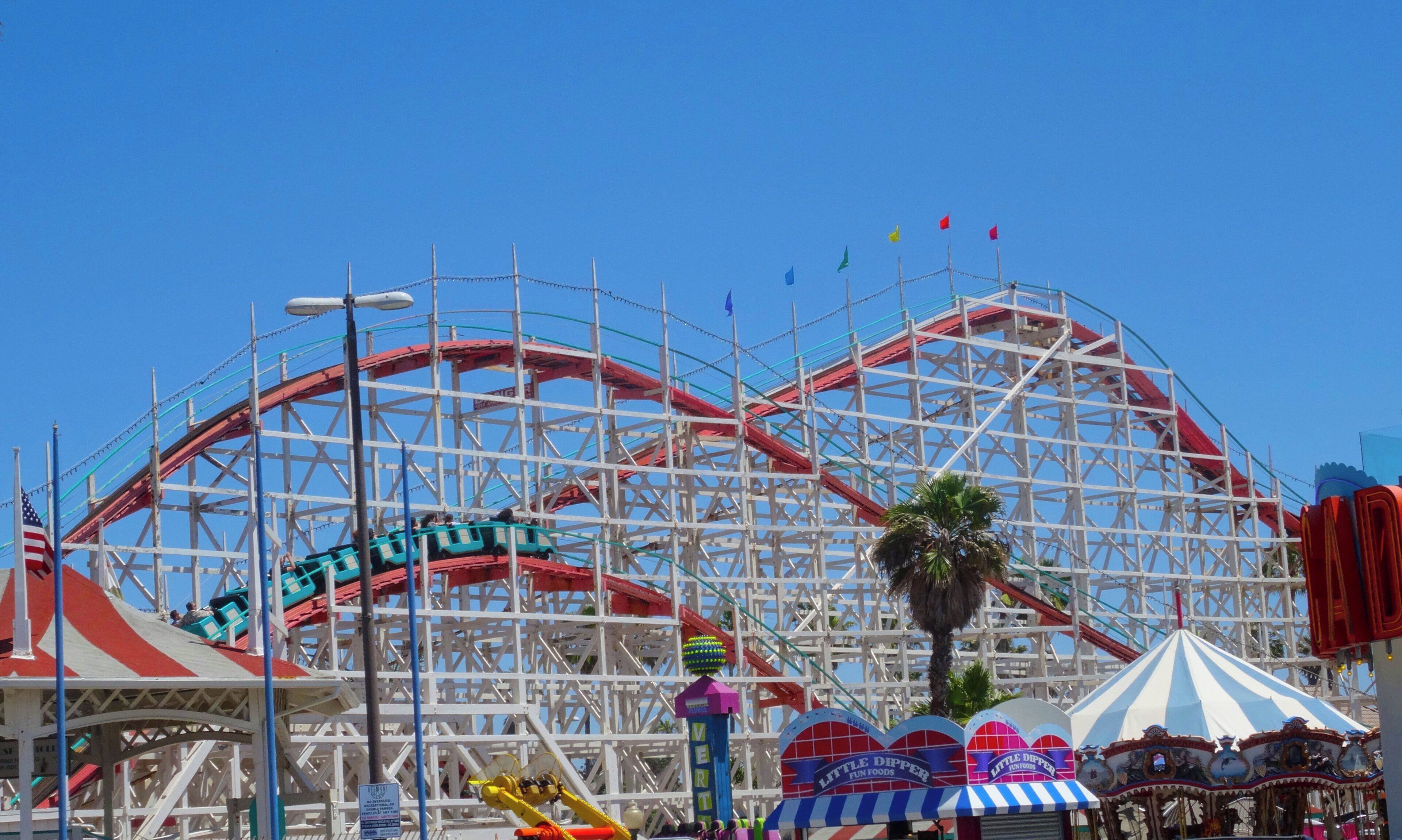 A roller coaster with a red track, surrounded by a white metal framework, and a carousel with striped canopies.