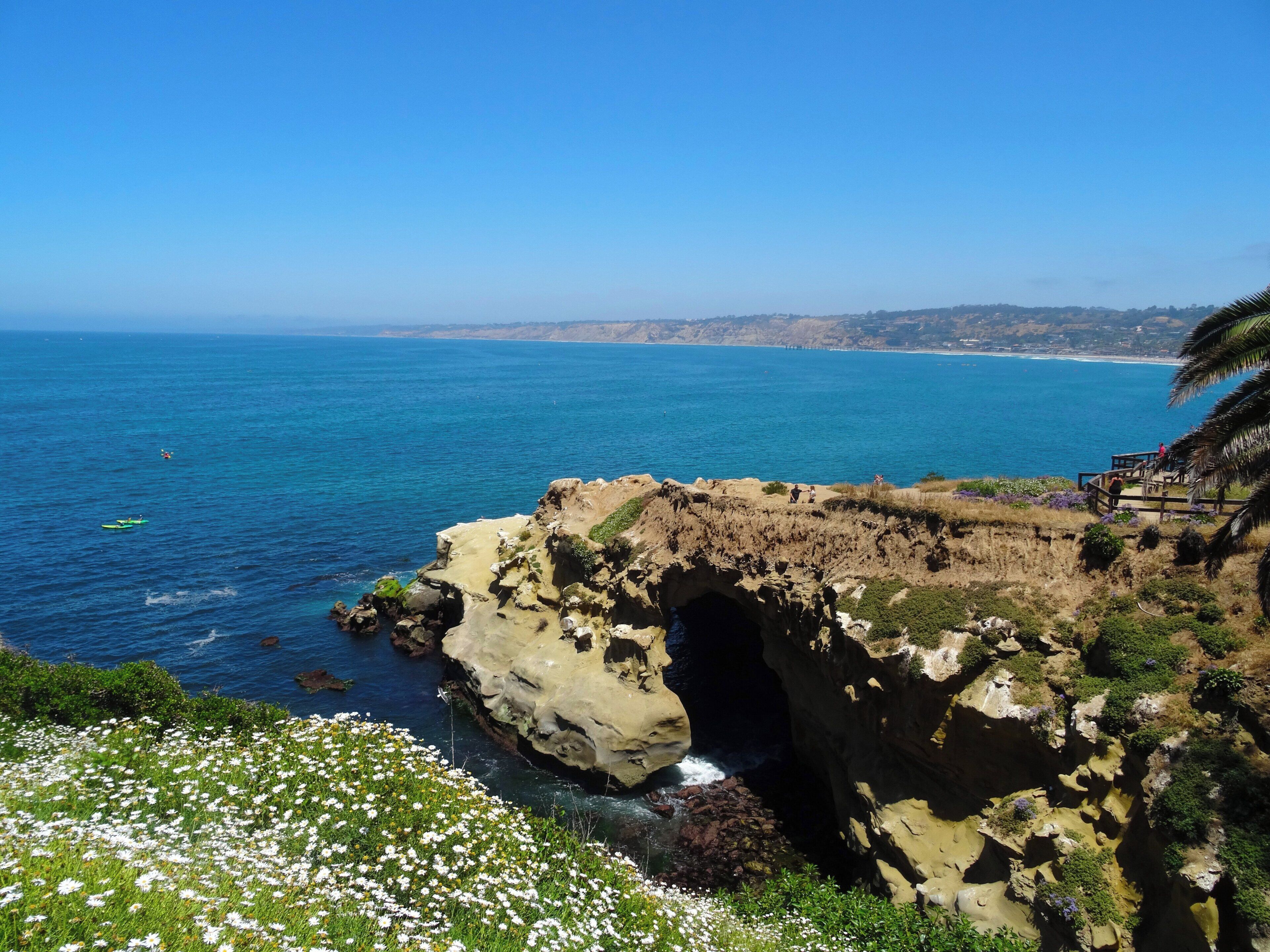A coastal landscape with a rocky cliff, a clear blue sea, and a pathway leading to a viewpoint overlooking the ocean.